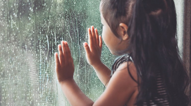 девочка смотрит на дождь сквозь стекло, девочка держит руки на стекле, за окном идет дождь