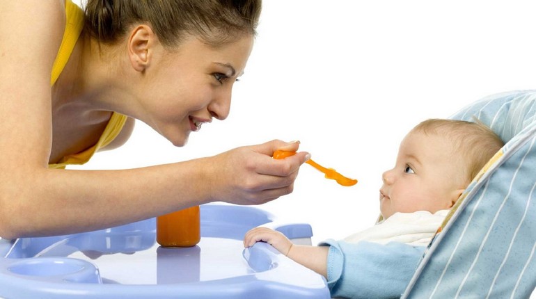 кормление малыша с ложечки, ребенок сидит в стульчике для кормления и мама кормит его с ложечки