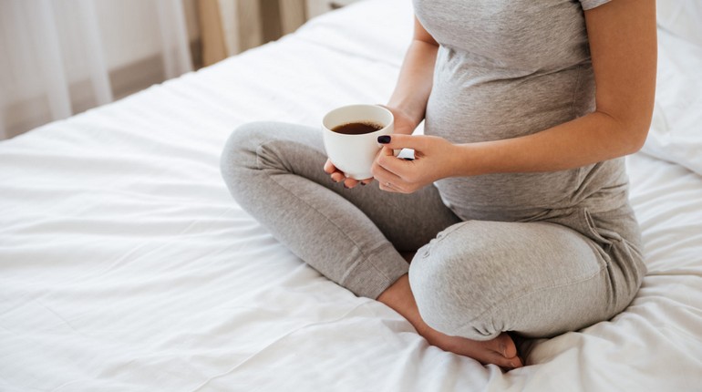 беременная девушка пьет кофе, девушка в положении сидит на кровати по-турецки с чашкой кофе в руках